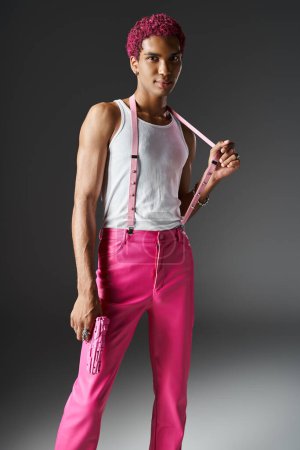 junger, stylischer Mann mit lockigem, rosa Haar, Hand auf Hosenträger, Spielzeugpistole in der Hand und Blick in die Kamera