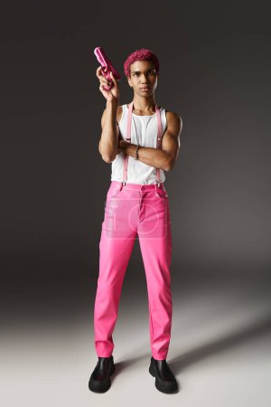 elegante hombre afroamericano con el pelo rosa apuntando a su pistola de juguete rosa posando sobre fondo gris