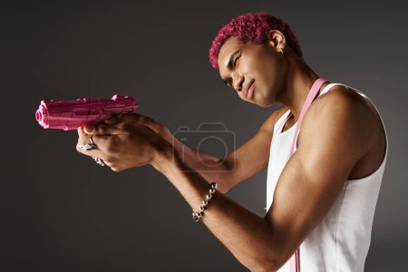 Téléchargez les photos : Rose cheveux afro-américain modèle masculin en pantalon avec des bretelles visant son pistolet jouet rose de côté - en image libre de droit