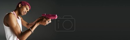 beau jeune modèle masculin en tenue vibrante avec des accessoires en argent visant son pistolet jouet rose loin