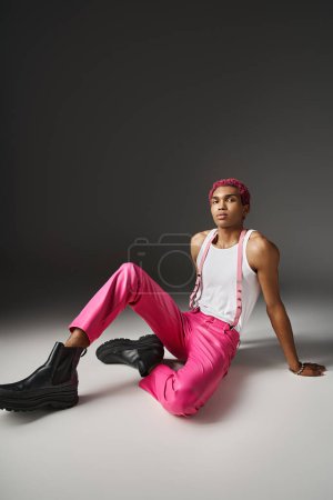 Foto de Hombre afroamericano bien parecido sentado en el suelo en pantalones de color rosa con tirantes, concepto de moda - Imagen libre de derechos