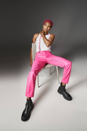 modelo masculino afroamericano en pantalones rosas con tirantes posando en silla blanca, concepto de moda