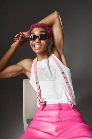 verspielter junger Mann mit rosa Haaren und Hosenträgern, der mit Sonnenbrille lächelt, Modekonzept