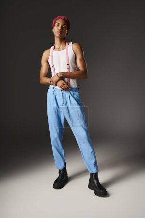elegante hombre afroamericano n pantalones azules con tirantes de color rosa mirando a la cámara, concepto de moda