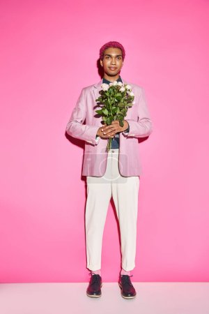 good looking pink haired man posing with rose bouquet on pink backdrop and smiling cheerfully