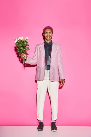 cheerful young man with bouquet of white roses posing on pink backdrop smiling sincerely at camera