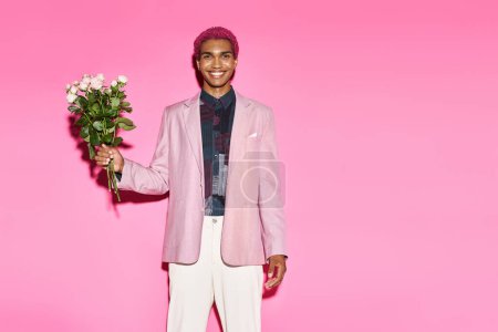 bel homme aux cheveux roses posant avec bouquet de roses sur fond rose souriant joyeusement à la caméra