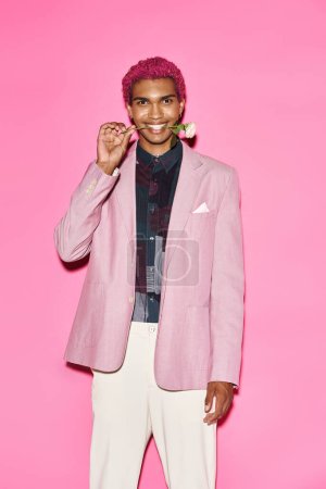 good looking cheerful man posing with rose in mouth on pink background and smiling at camera