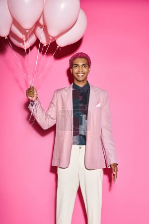 alegre modelo masculino sonriendo felizmente con globos en las manos y mirando a la cámara en el fondo rosa