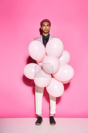 schöner Mann mit rosa Haaren hält rosa Luftballons in der Hand und blickt in die Kamera, die auf rosa Hintergrund posiert