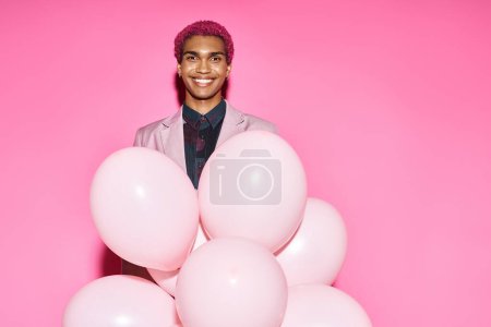 stylish african american man in pink blazer posing unnaturally with balloons on pink backdrop