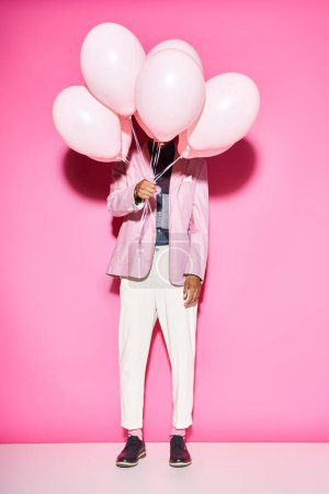 hombre con estilo en traje vibrante sosteniendo globos en frente de su cara posando sobre fondo rosa