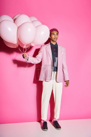 schöner Mann mit lockigem rosa Haar in rosa Blazer posiert mit Luftballons auf rosa Hintergrund, puppenhaft