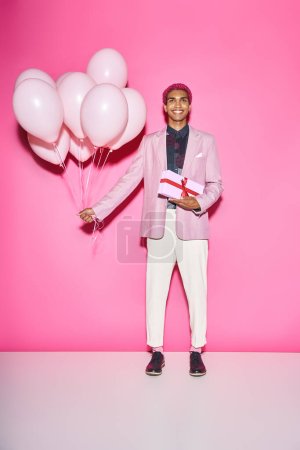 joyous young man in blazer posing with balloons and present smiling unnaturally on pink backdrop