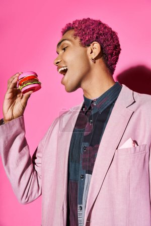 joyeux jeune homme en blazer rose avec des boucles d'oreilles en argent manger mini hamburger posant sur fond rose