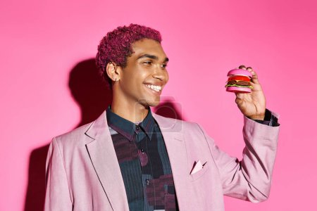 cheerful african american man smiling unnaturally and looking at mini burger on pink backdrop
