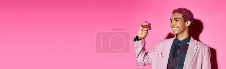 joyful handsome man with curly hair looking cheerfully at mini burger on pink background, banner