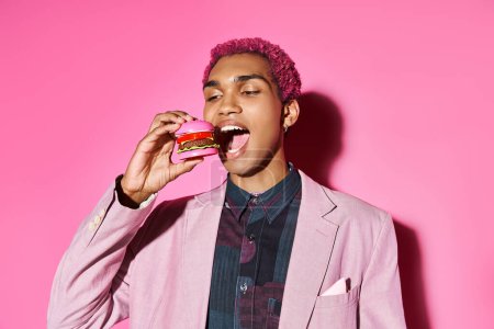 hombre afroamericano con estilo con pendientes de plata comiendo mini hamburguesa posando sobre fondo rosa