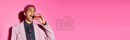 stilvoller gutaussehender Mann mit rosa gelockten Haaren und Ohrringen, der Mini-Burger auf rosa Hintergrund isst, Banner