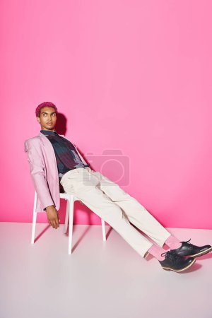 joven hombre guapo en chaqueta rosa con pantalones blancos sentado en la silla en el telón de fondo rosa, muñeca como
