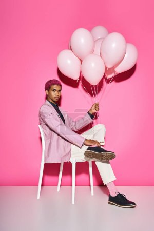 gut aussehender Mann mit rosa Haaren sitzt auf weißem Stuhl und hält Luftballons in die Kamera