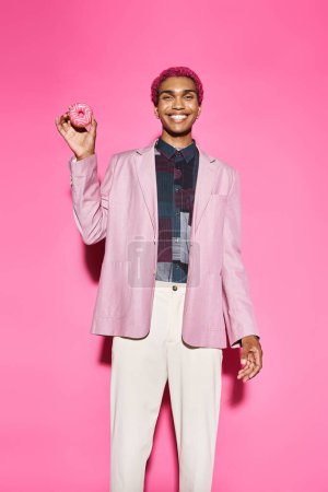 schöne afrikanisch-amerikanische männliche Modell posiert unnatürlich lächelnd und hält Donut, rosa Hintergrund