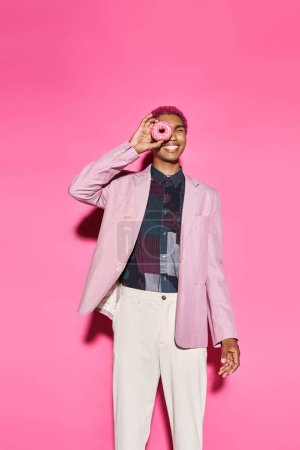 verspielter junger Mann mit lockigem rosa Haar posiert mit Donut in der Nähe seines Gesichts vor rosa Hintergrund