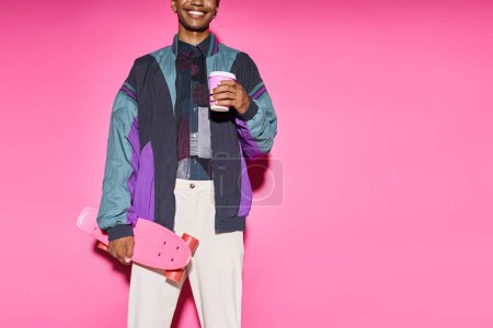 cropped view of cheerful young man in vibrant attire posing with coffee and skateboard, doll like