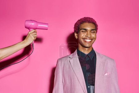 hombre guapo con el pelo rizado de color rosa posando con los ojos cerrados sobre fondo rosa, mano con secador de pelo