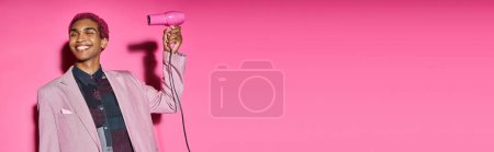 Foto de Elegante hombre de pelo rosa sonriendo extrañamente con los ojos cerrados con secador de pelo en las manos, pancarta - Imagen libre de derechos