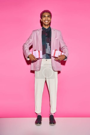 Photo for Good looking man with curly hair posing unnaturally and smiling with presents on pink backdrop - Royalty Free Image