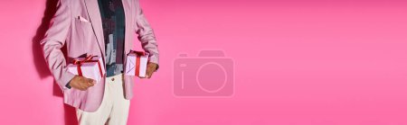 Foto de Vista recortada de hombre joven con estilo en traje vibrante con regalos en las manos sobre fondo rosa, pancarta - Imagen libre de derechos