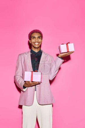 fröhliches junges männliches Model posiert mit Geschenken in der Hand vor rosa Hintergrund und wirkt wie eine Puppe