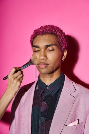 schöner Mann mit lockigem rosa Haar, der mit geschlossenen Augen posiert, Hand mit Schminkpinsel, puppenhaft