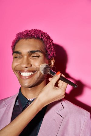 cheerful young man smiling sincerely with closed eyes, hand with makeup brush on pink backdrop