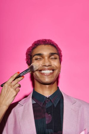 fröhliche stilvolle männliche Modell in lebendiger Kleidung lächelnd mit geschlossenen Augen, Hand mit Make-up-Pinsel