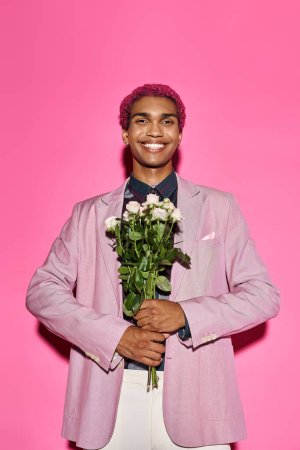 cheerful man with curly hair in pink blazer posing with rose bouquet and looking at camera