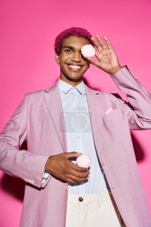cheerful young man in stylish outfit acting unnaturally with zefir in his hands on pink backdrop
