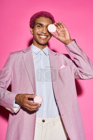 junger stylischer Mann lächelt unnatürlich und posiert mit Zefir in den Händen vor rosa Hintergrund