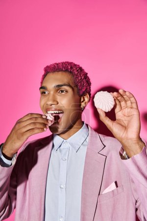fröhlicher junger Mann, der sich unnatürlich verhält, während er köstlichen rosa Zefir isst, der auf rosa Hintergrund posiert