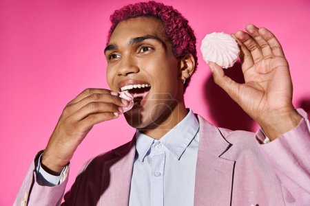 handsome pink haired man in pink blazer eating zefir posing unnaturally posing on pink backdrop
