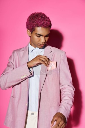 handsome man with curly pink hair in stylish attire on pink backdrop with present in his pocket
