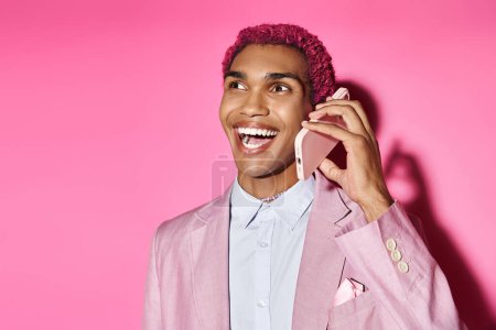 cheerful young african american male model in pink blazer posing with mobile phone on pink backdrop