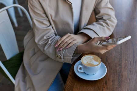Beschnittene junge Frau im Trenchcoat mit Smartphone in der Nähe von Tasse mit Cappuccino in Outdoor-Café