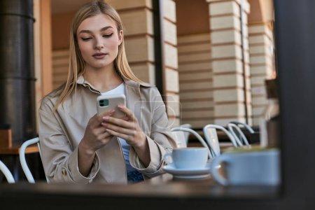 blonde young woman in trench coat using smartphone near cup of coffee while sitting in outdoor cafe