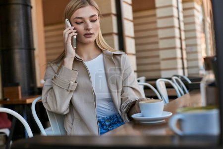 jolie jeune femme en trench coat ayant appel sur smartphone près de tasse avec cappuccino dans un café en plein air