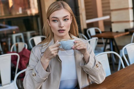 verträumte blonde Frau im Trenchcoat genießt ihre Tasse Cappuccino, während sie draußen im Café sitzt