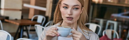 verträumte blonde Frau im Trenchcoat genießt ihre Tasse Cappuccino, während sie im Café sitzt, Banner