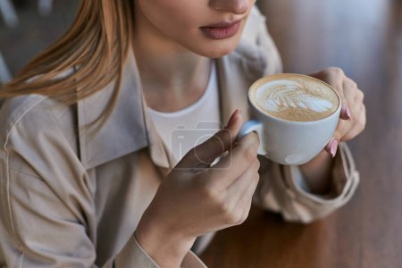 gestutzte blonde Frau im Trenchcoat genießt ihre Tasse Cappuccino im Café