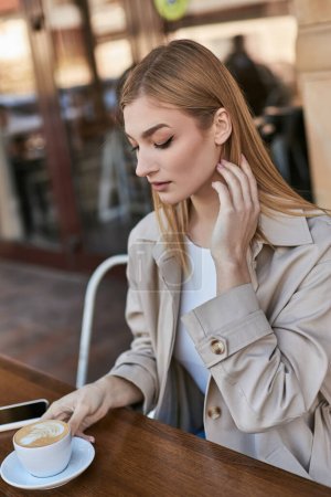 attraktive Frau im Trenchcoat genießt eine Tasse Cappuccino, während sie neben dem Smartphone im Café sitzt